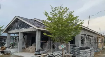 A RumahHQ bungalow at structural stage — gable steel trusses visible at the front, roof tiles beginning on the other bays, concrete columns and beams still unfinished, surrounded by scaffolding and stacked materials